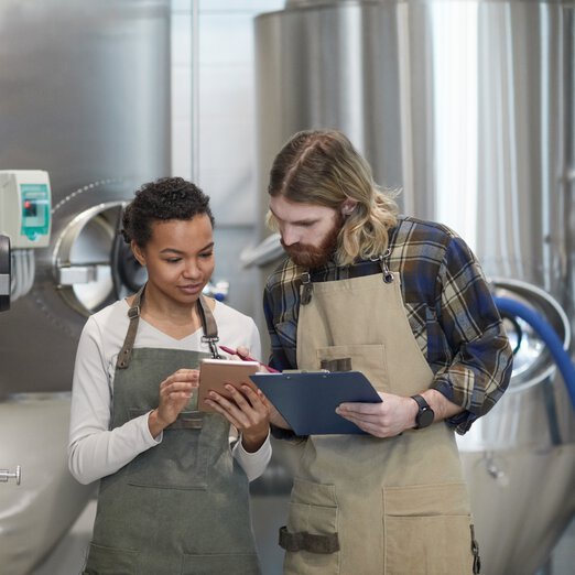 2 brewery employees with aprons talking in front of a brewing plant, bent over their tablets